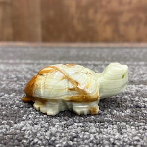 May include: A small, carved turtle figurine. The turtle has a light green body with brown and cream-coloured markings on its shell. The figurine is made of a stone-like material and has a detailed, textured appearance.