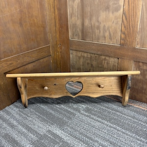 Puede incluir: Estante de madera con un recorte en forma de corazón y dos pequeñas clavijas de madera. El estante tiene un acabado de madera natural y un borde festoneado. El estante mide aproximadamente 61 cm de largo.