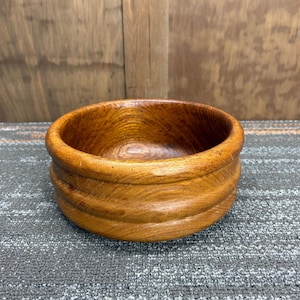 May include: A handcrafted wooden bowl with a warm, reddish-brown hue. The bowl features a rounded shape with a decorative, ridged band around the exterior. The wood grain is visible, adding to its natural aesthetic.