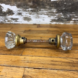 May include: Antique glass door knobs with brass hardware. The clear glass knobs have a ribbed and rounded design. The connecting rod is rusty, showing the age of the hardware. The door knobs are on a wooden surface.