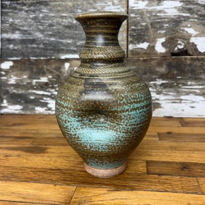 May include: A handcrafted ceramic vase with a textured surface. The vase features a brown and turquoise color scheme with a rope-like design around the neck. The base has a natural, unglazed finish. This decorative vase is suitable for home decor.