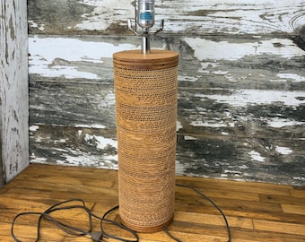 May include: A cylindrical table lamp with a brown, textured shade. The lamp has a silver neck and a light bulb socket. The base and top of the lamp are made of wood. The lamp is plugged into an electrical outlet.