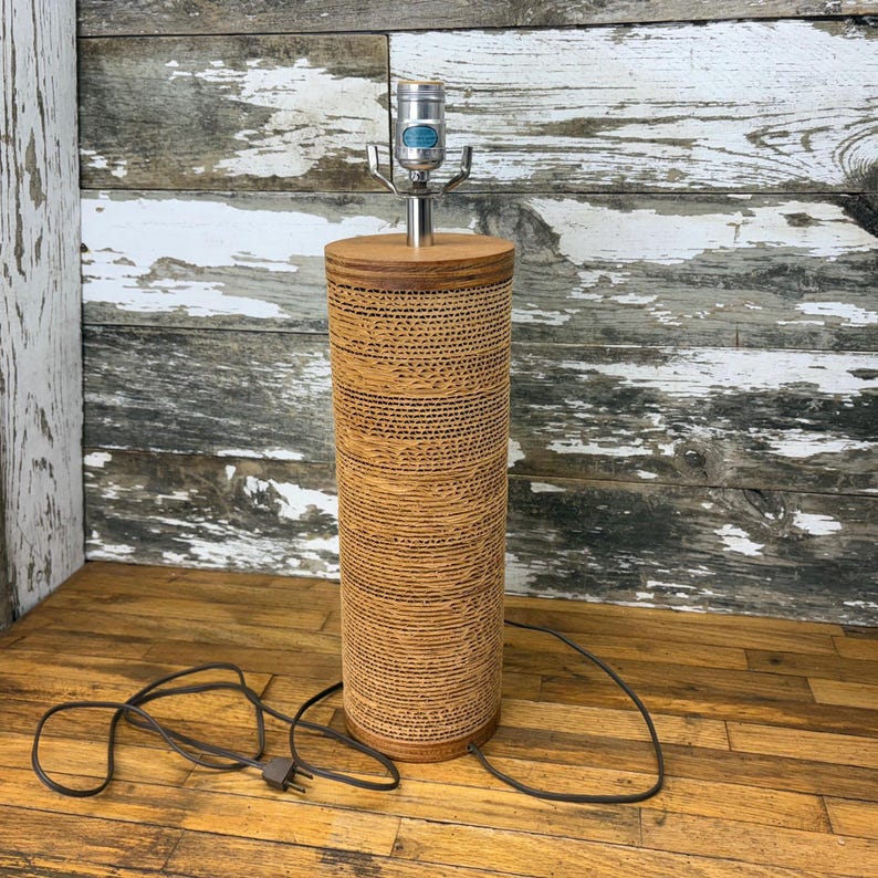 May include: A cylindrical table lamp with a brown, textured shade. The lamp has a silver neck and a light bulb socket. The base and top of the lamp are made of wood. The lamp is plugged into an electrical outlet.