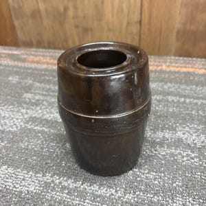 Puede incluir: Un jarrón de cerámica marrón oscuro de forma cilíndrica, ligeramente ensanchado en la base. El jarrón tiene un acabado liso y brillante y una abertura circular en la parte superior. Probablemente se utiliza para almacenamiento o decoración.