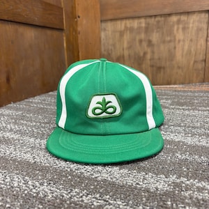 Puede incluir: Una gorra de béisbol de color verde brillante con dos franjas verticales blancas en los paneles frontales. Centrado en la parte delantera hay un parche blanco con un logotipo bordado en verde que representa una planta estilizada.