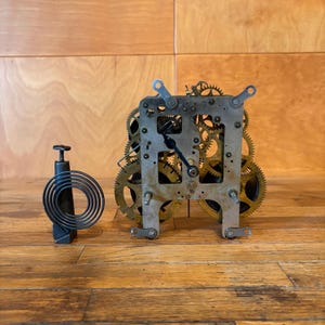 May include: A mechanical clock movement featuring a complex arrangement of brass gears and a silver-toned metal frame. Beside the mechanism stands a separate black metal component with a coiled spring attached to a vertical post and base.