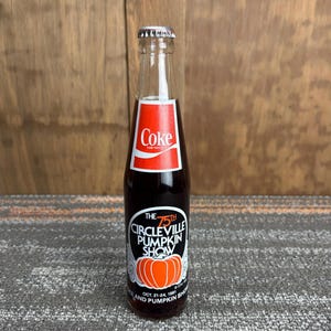 May include: A vintage Coke bottle with a red and white label. The bottle features a black and white label for the 75th Circleville Pumpkin Show, with an orange pumpkin graphic. The bottle is clear with a silver cap.