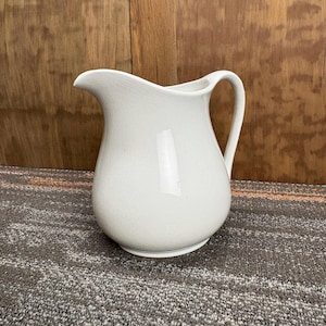 May include: A white ceramic pitcher with a curved spout and a handle. The pitcher has a rounded body and sits on a flat base. The surface has a slightly textured appearance. The background is a wooden surface.