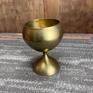 May include: A brass-colored goblet with a rounded bowl and a pedestal base. The bowl has a slight tilt, and the goblet has a smooth, polished finish. The stem is a smooth, rounded shape. The goblet is sitting on a gray and white patterned surface.