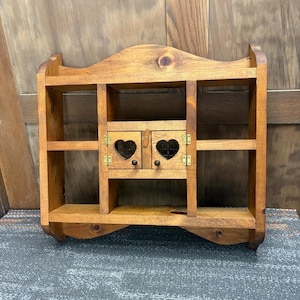 May include: A medium-brown wooden wall shelf featuring an arched top, two open side compartments, and a central cabinet with two small doors. Each door includes a heart-shaped cutout, a small round knob, and gold-toned metal hinges.