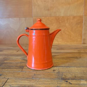 May include: A bright orange enamel coffee pot with a matching lid, a curved handle, and a tapered spout. The pot features a smooth, glossy finish with a black rim detail around the base and the lid, showcasing a classic cylindrical design.