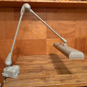 May include: A light gray adjustable desk lamp with a long, articulated arm and a rectangular, horizontal light head. The lamp features a sturdy clamp base for mounting to a surface and includes a small, dark power switch on the top housing.