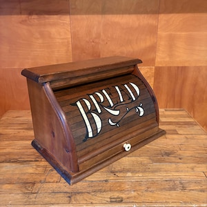 Peut inclure: Une boîte à pain en bois avec un couvercle coulissant sur lequel est peint le mot BREAD en blanc avec des contours noirs. Le récipient présente une finition en bois brun moyen et un petit bouton blanc sur le panneau avant pour ouvrir le couvercle.