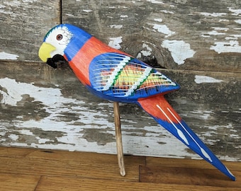 May include: A hand-painted wooden parrot figurine. The bird has a blue head, red body, and blue wings with yellow, green, and white accents. The parrot is mounted on a wooden dowel, set against a weathered wood background.