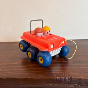 Puede incluir: Un coche de juguete rojo vintage con ruedas azules y detalles amarillos. El coche tiene dos figuras sonrientes dentro y las palabras "Bouncing Buggy" impresas en el lateral. Una cuerda amarilla está adjunta.