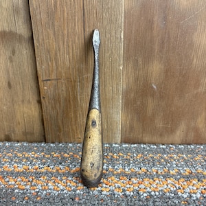 May include: A vintage flathead screwdriver with a wooden handle. The metal blade is silver and shows signs of wear. The handle is a light brown color with a few dark spots. The tool is set against a wooden background.