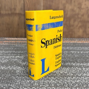 May include: A yellow Langenscheidt Pocket Spanish Dictionary. The book has blue accents and text that reads "Spanish Dictionary" and "Spanish English". The book is standing upright.