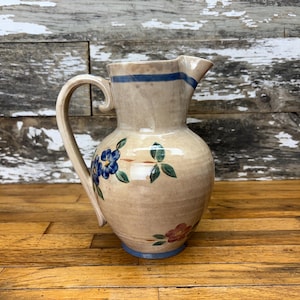 May include: A beige ceramic pitcher with a curved handle and spout. The pitcher is decorated with blue floral and green leaf designs, with a blue stripe around the top and base. The pitcher is sitting on a wooden surface.