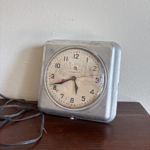 May include: A vintage square-faced clock with a silver metal casing. The clock face is off-white with black numerals and red accents. The clock hands are black and red. A black power cord extends from the clock.