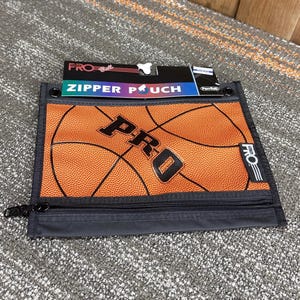 May include: Orange basketball-themed zipper pouch with the word "PRO" in black. The pouch has a black zipper and a tag that reads "ZIPPER POUCH". The background is a neutral gray.