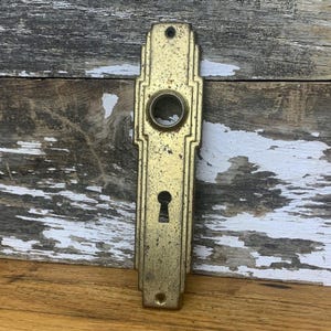 May include: A vintage brass door plate with an Art Deco design. The plate has a keyhole and a round hole for a knob. The plate is slightly tarnished, giving it a rustic look.