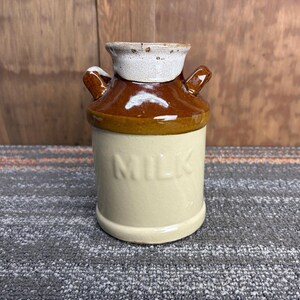 May include: A vintage ceramic milk jug with a cream-colored base and a brown top. The word "MILK" is embossed on the front. It has two small handles and a white rim. The jug is likely used for serving milk.