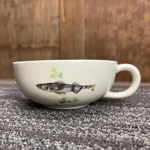 May include: A white ceramic soup bowl with a handle, featuring a detailed illustration of a fish and aquatic plants. The fish is primarily black and white with yellow accents. The bowl is on a gray surface.