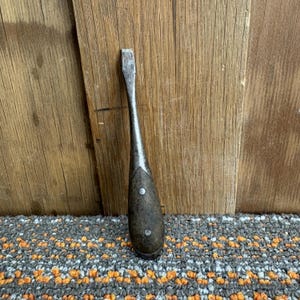 May include: A vintage flathead screwdriver with a dark wooden handle and a metal blade. The handle is secured with two small silver rivets. The tool is set against a wooden background and a patterned rug.