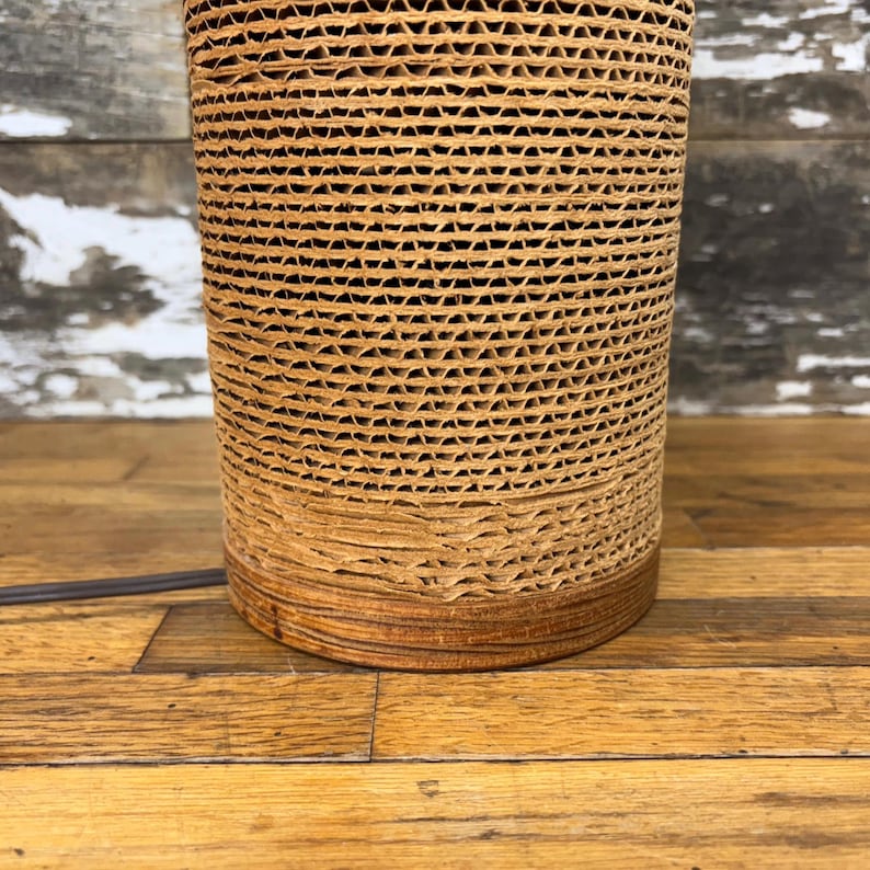 May include: A cylindrical lamp base made from corrugated cardboard. The lamp has a natural, light brown colour with a darker brown base. A black power cord extends from the base. The lamp sits on a wooden surface.