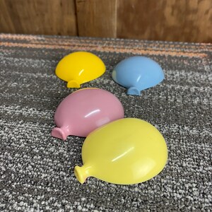 May include: Four colorful plastic balloon-shaped toys in yellow, pink, and blue. The toys are on a gray and white patterned surface. Each balloon has a small, rounded tail.