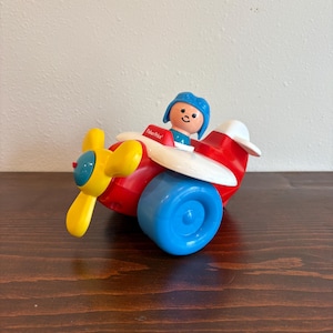 May include: A vintage Fisher-Price toy airplane. The red plane has a yellow propeller, blue wheels, and a white wing. A small figure with a blue hat sits in the cockpit. The toy is on a dark brown wooden surface.