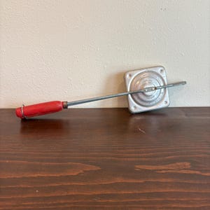 Puede incluir: Un sándwichera de metal vintage con un mango de madera rojo. La superficie de cocción cuadrada es de color plateado y tiene un diseño circular en el centro. El mango está unido a la superficie de cocción con una varilla metálica.