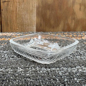 May include: A clear, square-shaped glass dish with a decorative etched pattern. The dish has a diamond-like shape and is sitting on a gray and orange patterned surface. The dish is empty.