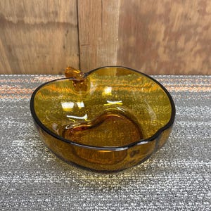 Puede incluir: Cuenco de vidrio color ámbar con forma de manzana y tallo. El cuenco tiene un borde oscuro y una hendidura en forma de corazón en el interior. El vidrio es transparente. Una pieza decorativa para servir o exhibir.