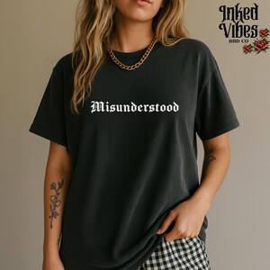 May include: Dark gray t-shirt with the word "Misunderstood" in white gothic font. The shirt has short sleeves and a relaxed fit. The brand logo "Inked Vibes and Co" is visible in the upper right corner.