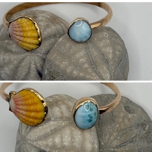 May include: A gold-toned open cuff bracelet featuring a yellow and pink scallop shell on one end and a smooth, light blue oval stone on the other. The metal band has a hammered texture, and the design is presented in two different angles.