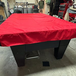 May include: A red pool table cover with black trim. The cover is made of a thick, durable fabric and is designed to protect the table from dust and dirt.