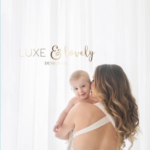K&ouml;nnte beinhalten: Eine Frau in einem wei&szlig;en Kleid h&auml;lt ein Baby in ihren Armen. Das Baby schaut in die Kamera und l&auml;chelt. Die Frau hat lange, lockige Haare. Der Hintergrund ist ein wei&szlig;er Vorhang. Der Text "LUXE & lovely DESIGN CO." ist im Bild sichtbar.