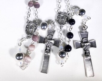 May include: Two silver rosaries with different coloured beads. One rosary has purple and grey beads, the other has dark blue beads. Both rosaries have silver crosses and a silver charm.