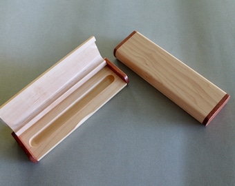 May include: A wooden pen box, open and closed, with a light wood body and contrasting dark wood trim. The open box reveals a carved interior designed to hold writing instruments. The box is rectangular and has a hinged lid.
