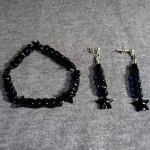 Puede incluir: Un brazalete negro con cuentas en forma de estrella y pendientes a juego. Las cuentas son de color azul oscuro y tienen un acabado brillante.