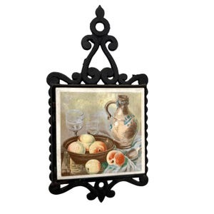 Puede incluir: Salvamanteles de hierro fundido negro con un marco decorativo y una inserción de baldosas de cerámica. La baldosa presenta una pintura de naturaleza muerta de melocotones, una jarra y vasos. El salvamanteles está diseñado para proteger las superficies de los utensilios de cocina calientes.