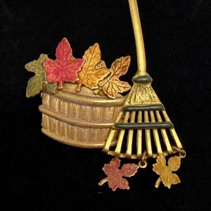 Puede incluir: Un broche de metal dorado que presenta una cesta llena de hojas de otoño pintadas en tonos rojo, naranja y verde. Un rastrillo de jardín está colocado junto a la cesta, con dos pequeños dijes de hojas colgando en la parte inferior.