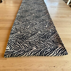 May include: A long, rectangular table runner featuring a repeating black and cream-colored zebra stripe pattern. The fabric has a textured weave and finished edges, extending lengthwise across the center of a light-toned wooden surface.