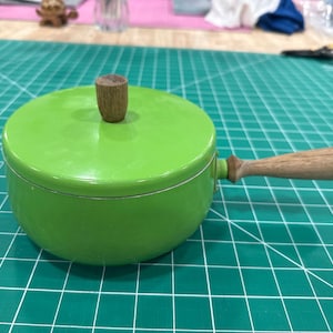 Puede incluir: Una pequeña cacerola verde brillante con tapa a juego y asa de madera. La tapa tiene un pequeño pomo cilíndrico de madera. La cacerola está sobre una superficie verde con patrón de cuadrícula.