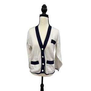 Puede incluir: Cárdigan color crema con ribetes y detalles en azul marino. El suéter de manga larga presenta un cuello en V, tres botones blancos y dos bolsillos delanteros. El ribete está alrededor del cuello, los bolsillos y los puños. El cárdigan se muestra en un maniquí.