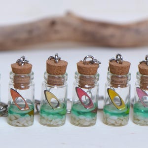 Op de afbeelding: Miniatuur glazen fles sleutelhangers met kurken stoppers. Elke fles bevat een kleurrijke miniatuur surfboard en decoratieve stenen. De flessen hebben een zilveren ketting en ring. De flessen zijn gevuld met groene en witte elementen.