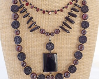 May include: A multi-strand necklace featuring black and burgundy beads. The necklace includes carved rose-shaped beads, black onyx beads, and a large black onyx pendant.