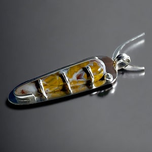 Peut inclure: Pendentif artisanal avec une pierre précieuse oblongue aux motifs jaunes, bruns et blancs. La pierre est sertie dans un cadre en métal argenté avec trois barres verticales et une boucle pour une chaîne. Le pendentif est présenté sur une surface réfléchissante.