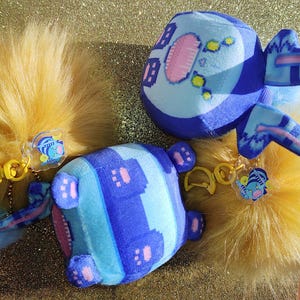 May include: Close-up of two plush toys with blue and light blue striped patterns, featuring pink accents and attached to yellow fur keychains. Each keychain has a small, cartoon-style charm. The background is a glittery gold.